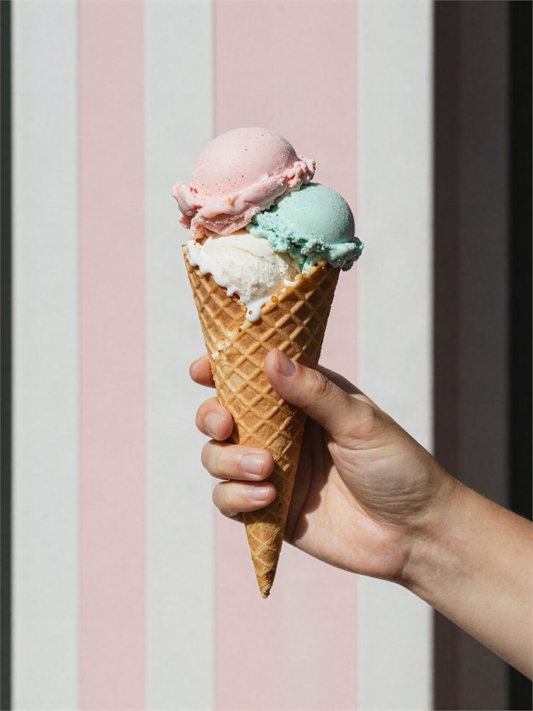 A two-scoop waffle cone held up against the pink and white striped wall.